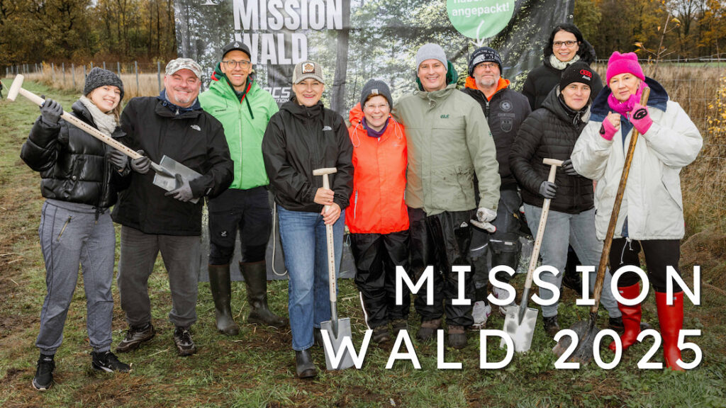 Gruppe von freiwilligen Helfern der Mission-Wald-Baumpflanzaktion steht mit Spaten auf einer vorbereiteten Pflanzfläche; sie lächeln in die Kamera vor einem Banner mit dem Schriftzug ‚Mission Wald‘